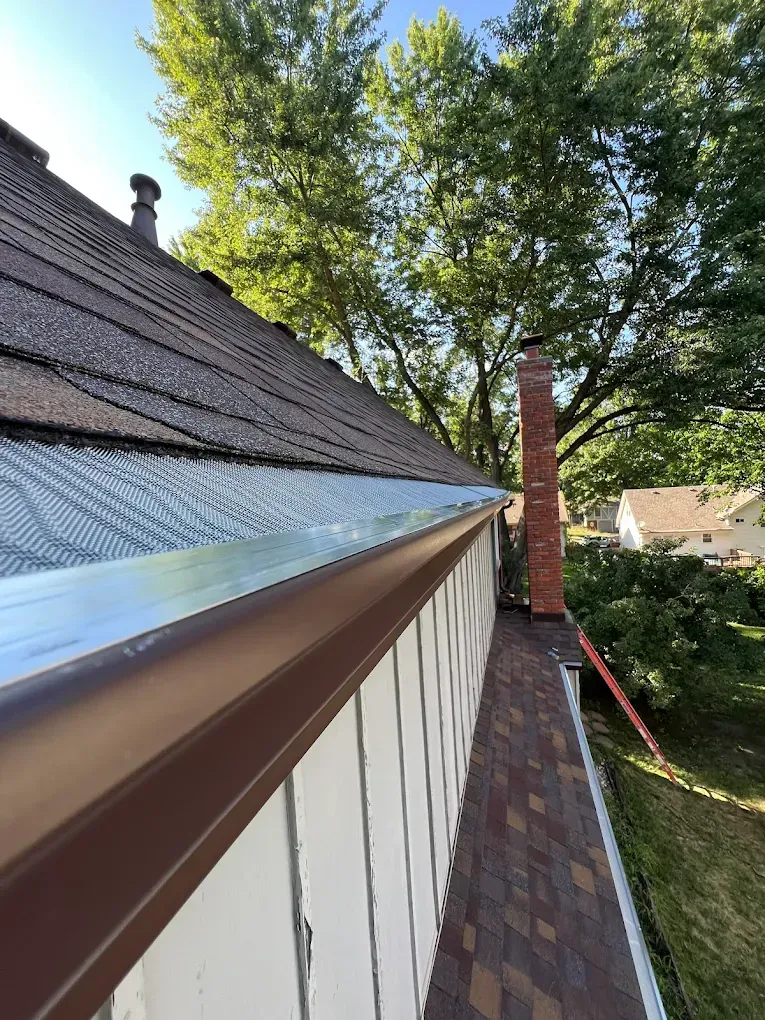 A brown gutter with a wire screen guard installed along the edge of a shingled roof next to a brick chimney.