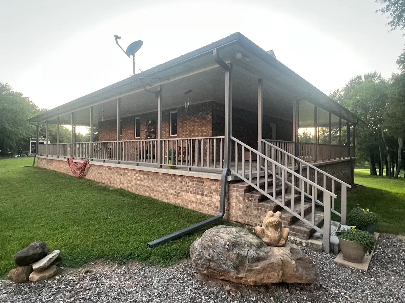 A brick house with a wrap-around porch and staircase, surrounded by a lawn, trees, and a large decorative stone.