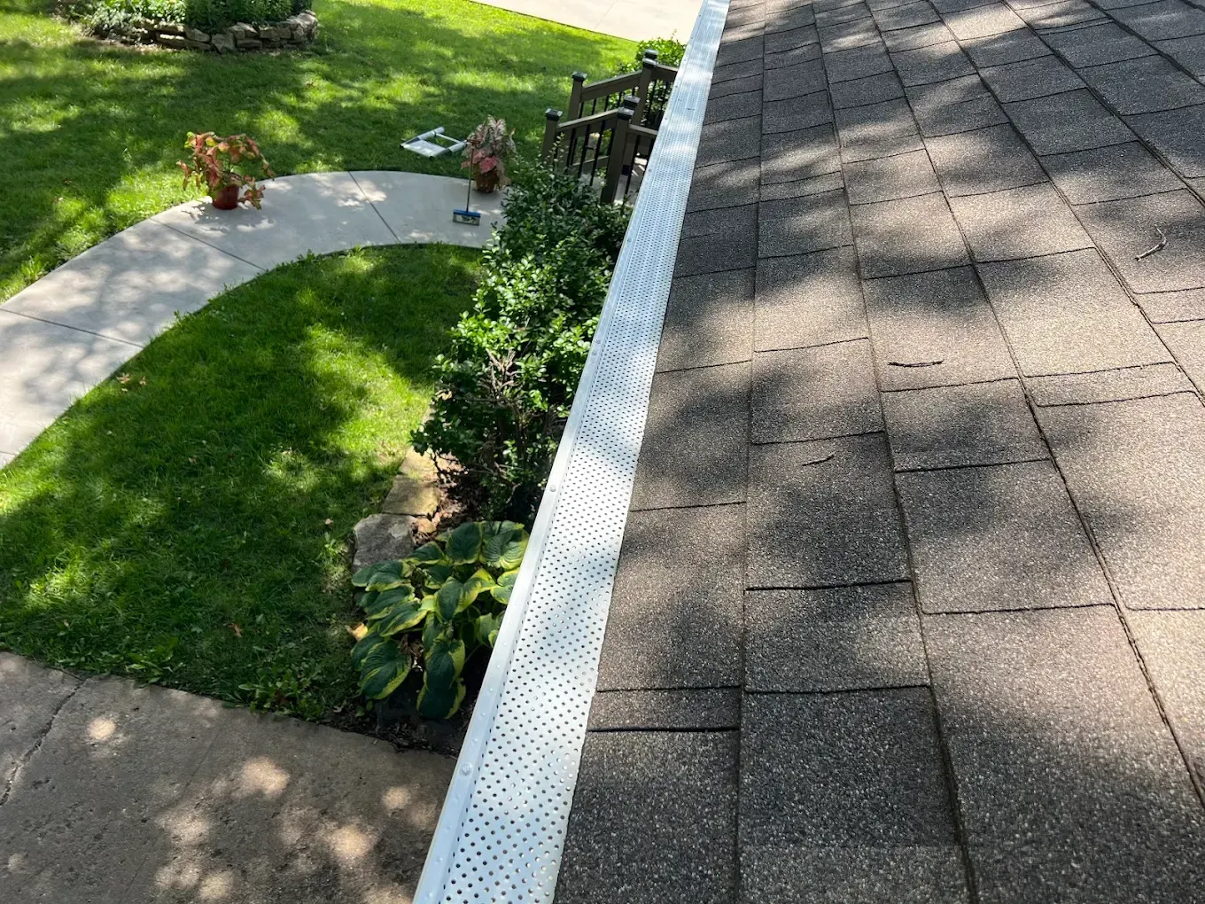 A view looking down from a shingled roof edge at a white gutter guard, overlooking a garden path and green lawn.