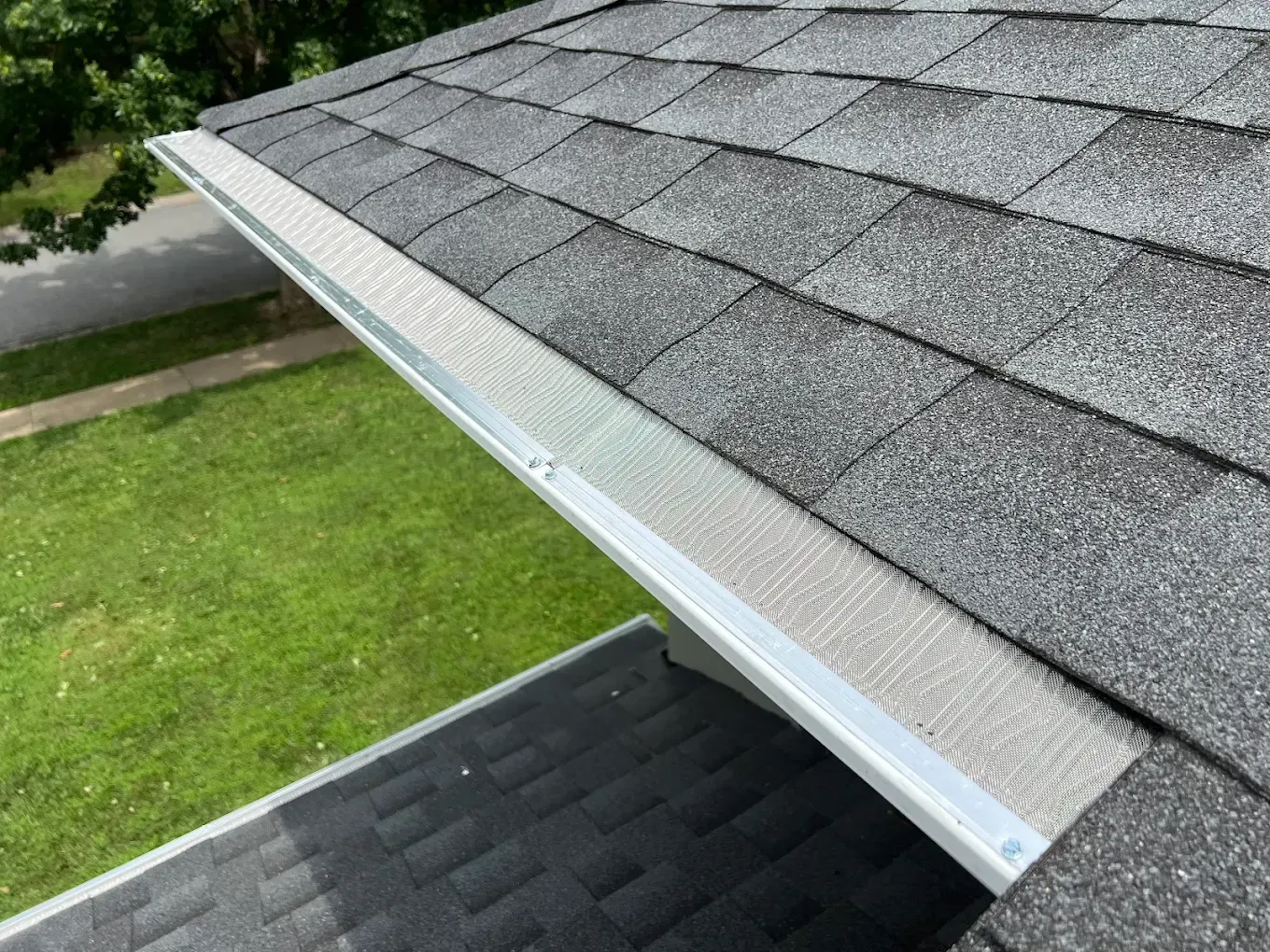 A section of a house roof featuring grey asphalt shingles and a metal gutter protected by a mesh gutter guard.