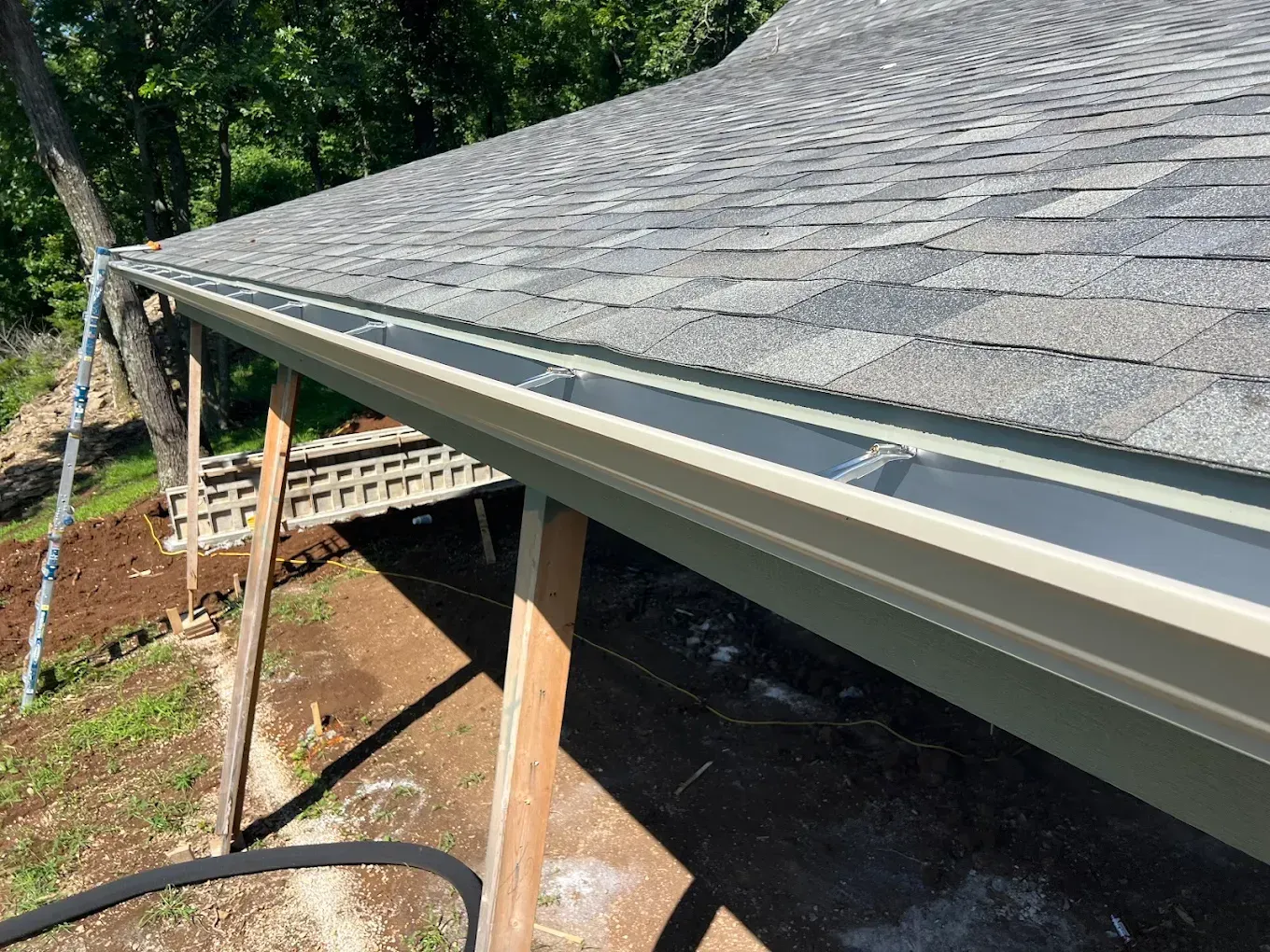 Tan seamless rain gutters installed along the edge of a grey asphalt shingle roof above a dirt construction site.
