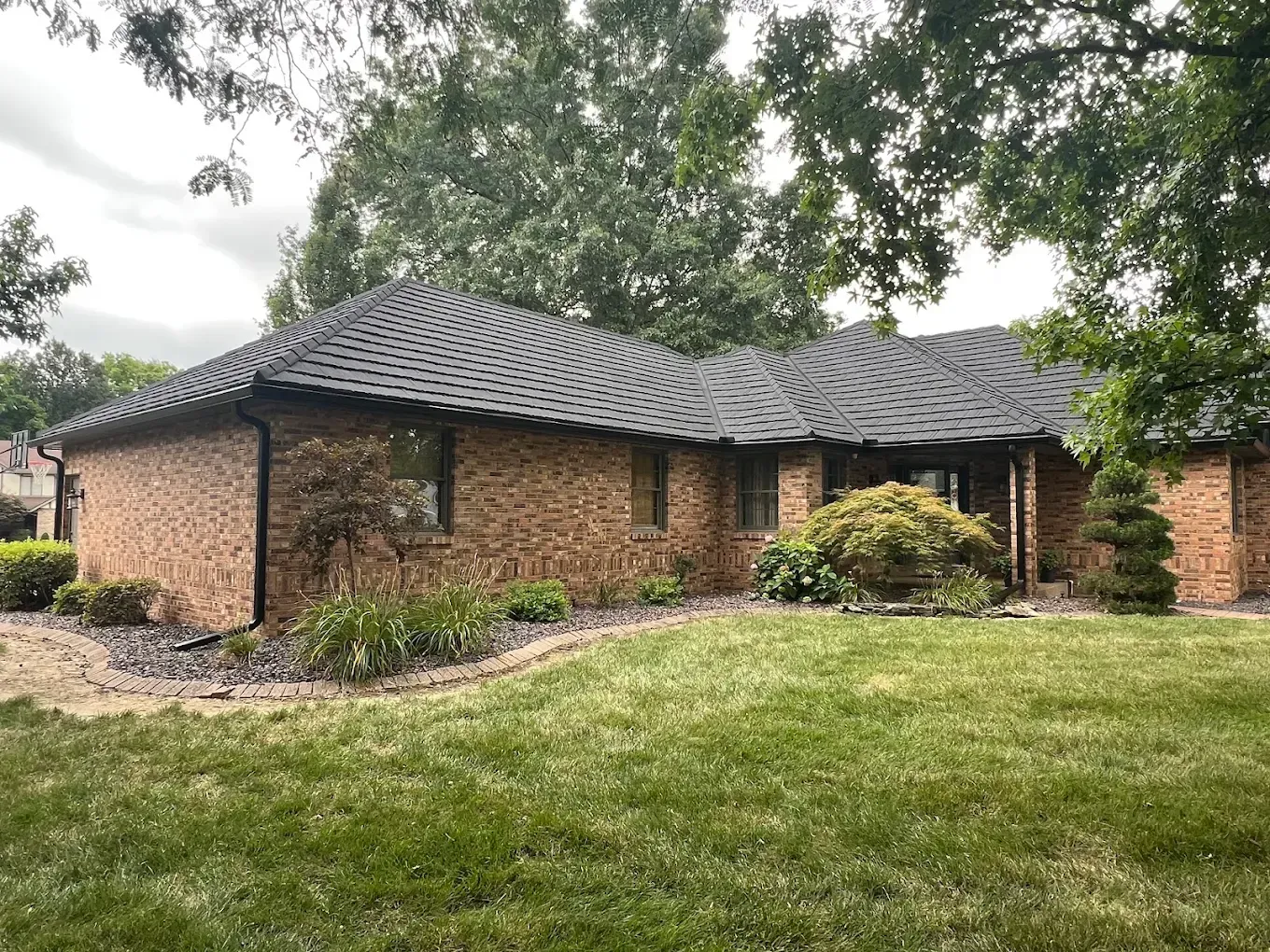 A single-story brick house with a dark shingled roof, surrounded by trees and a green lawn on an overcast day.