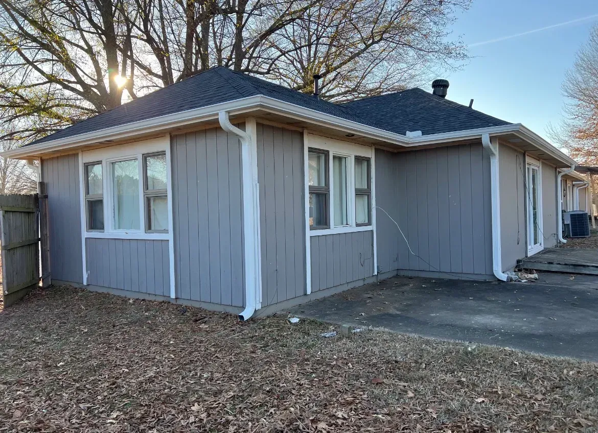A gray, single-story house with vertical siding and white trim under a dark shingled roof, set against a backdrop of trees.