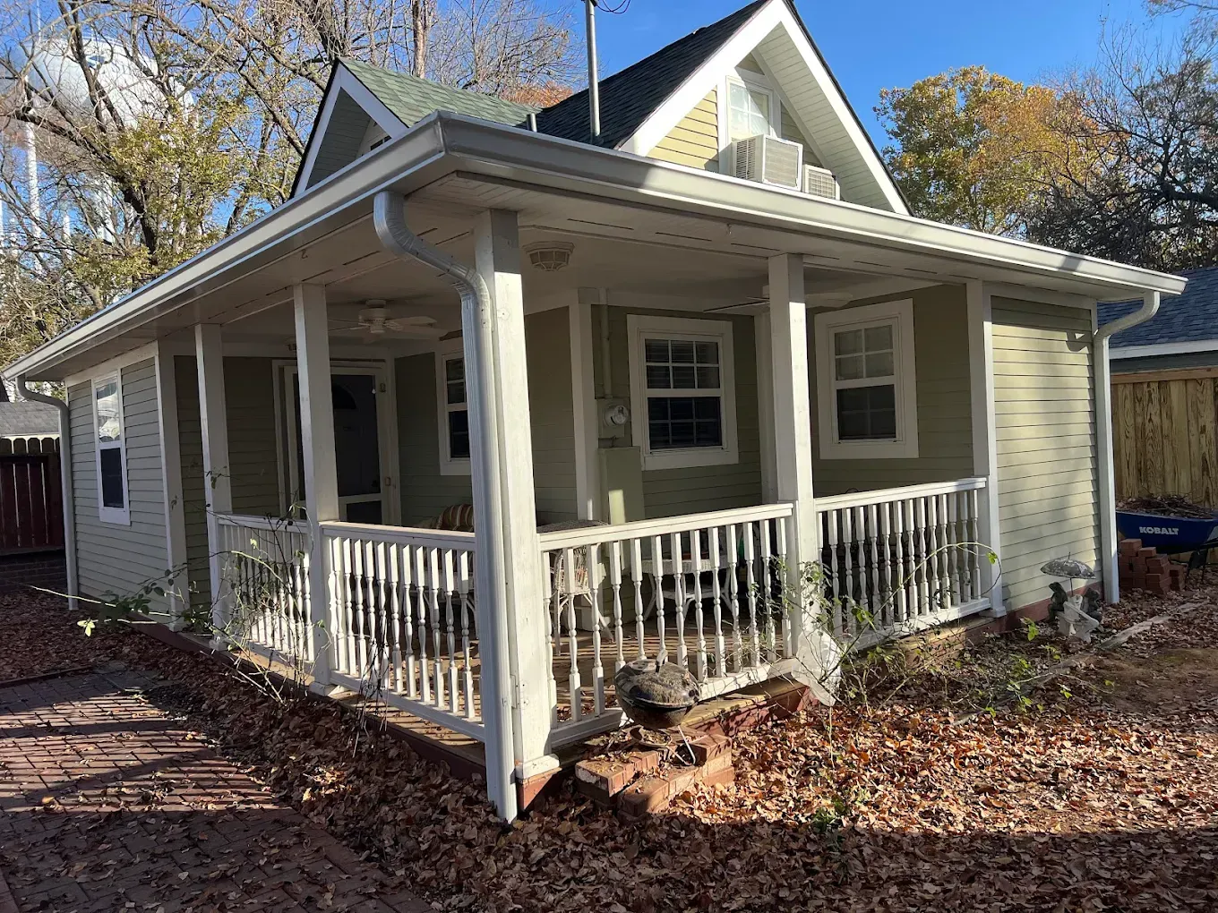 A sage green cottage with white trim and a wrap-around porch, surrounded by fallen autumn leaves.