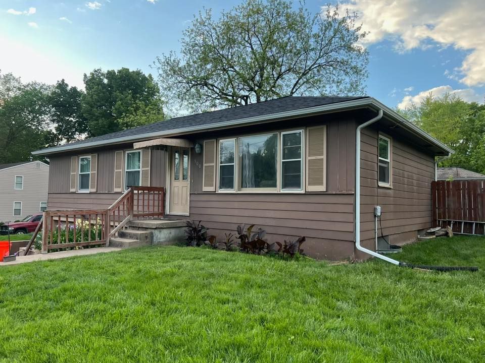 A one-story house with dark brown horizontal siding, tan shutters, a front porch, and a lush green lawn.