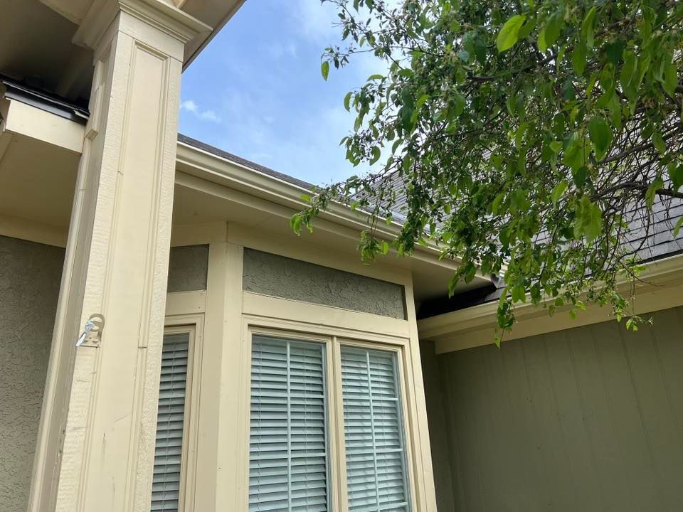 Low-angle view of a cream-colored house exterior with columns, windows with blinds, and green tree branches overhead.