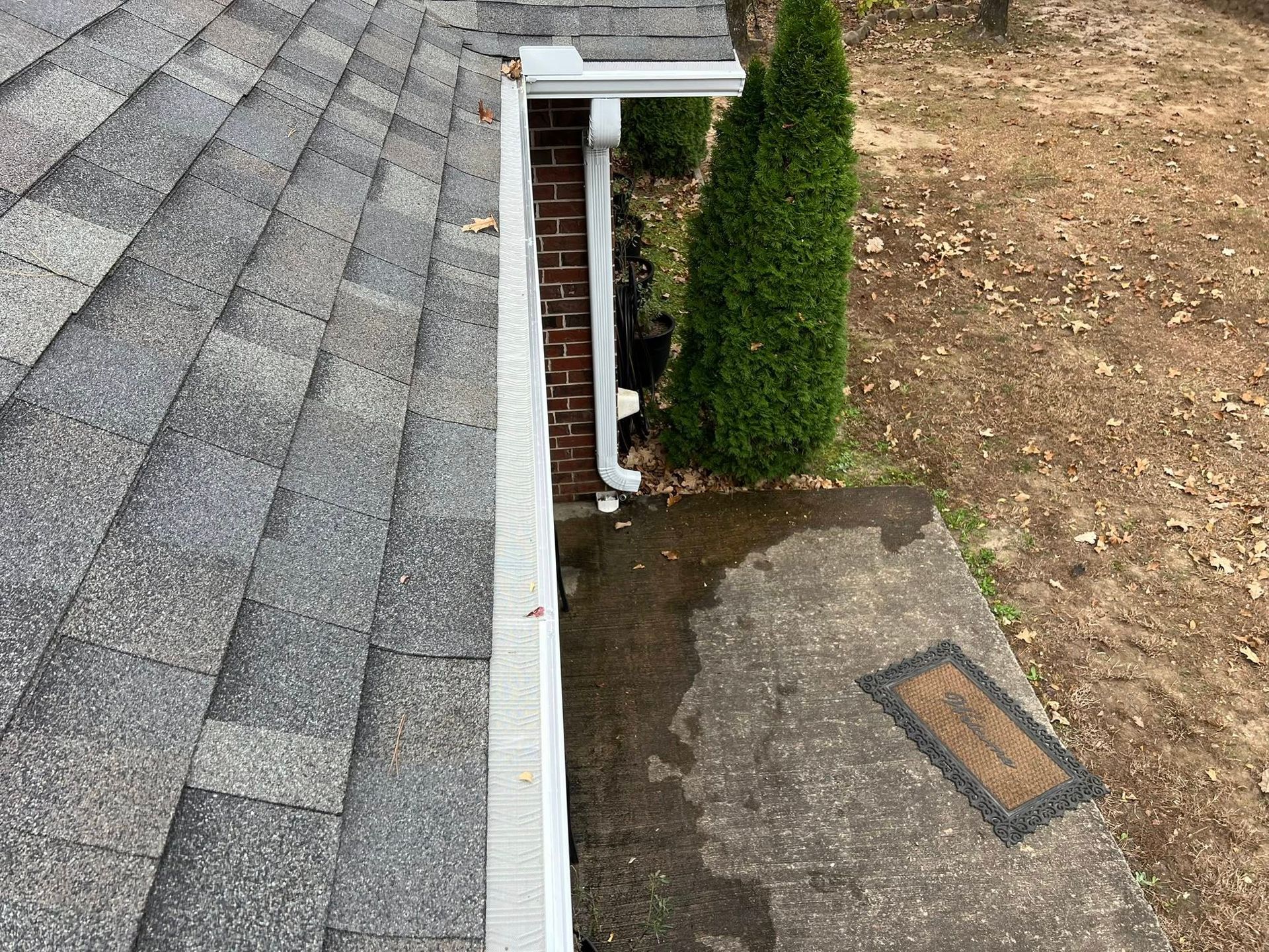 A high-angle view of a gray shingled roof edge, a white gutter downspout, and a damp concrete walkway with a small mat.