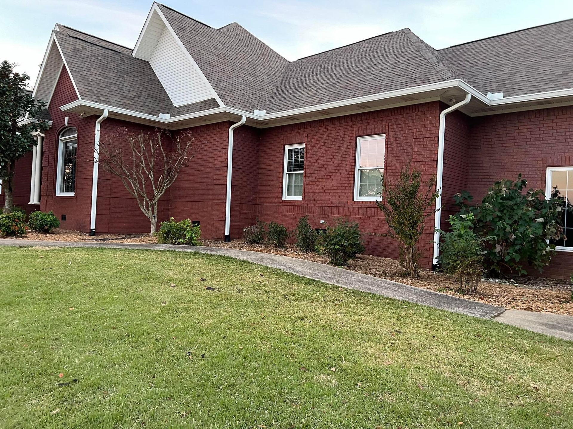 A single-story brick house with a dark red exterior, gabled roof, and a green lawn under a bright, cloudy sky.