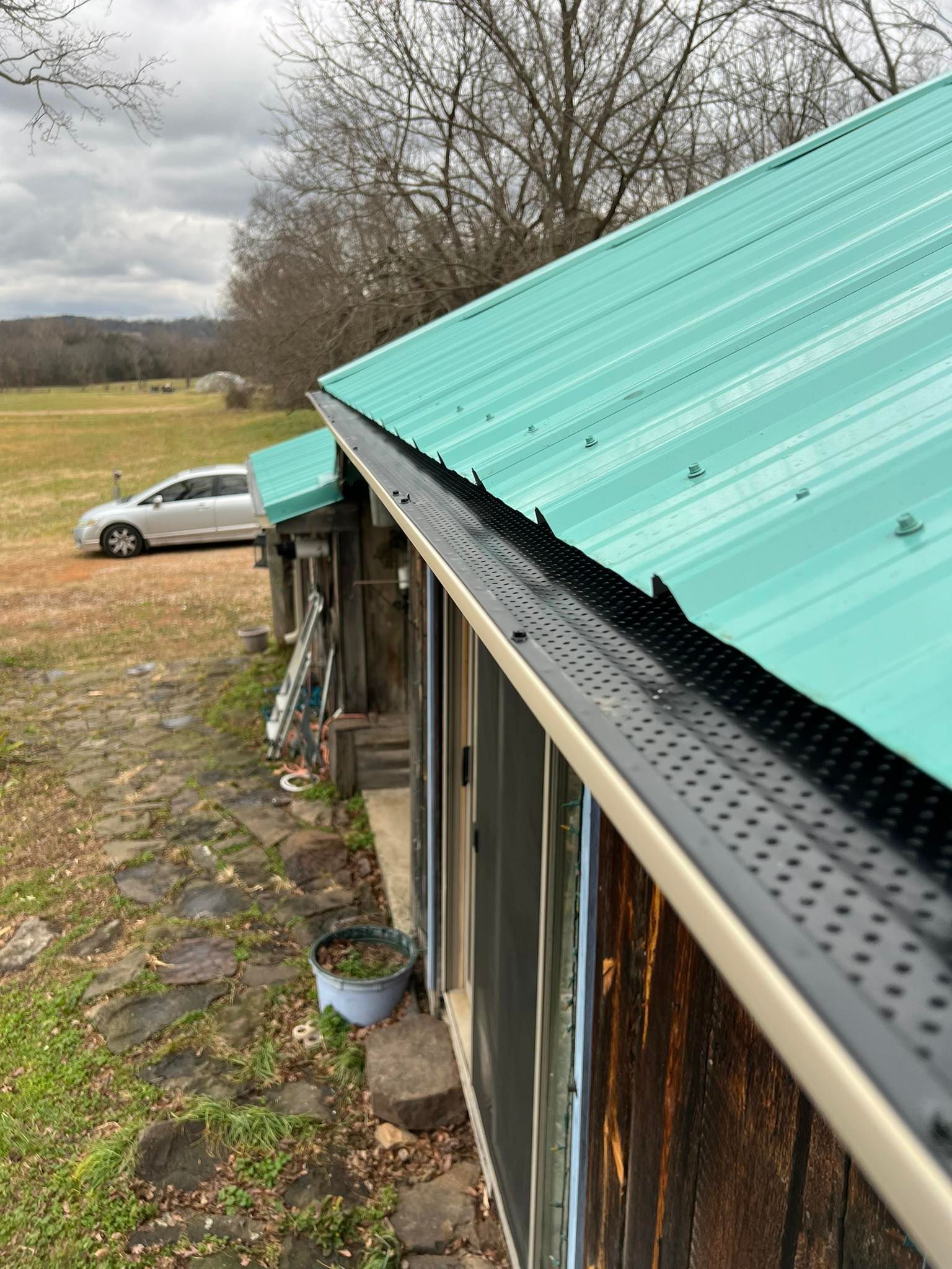 A view along a metal roof with a turquoise-colored paneled surface and a black gutter guard, looking out to a field.