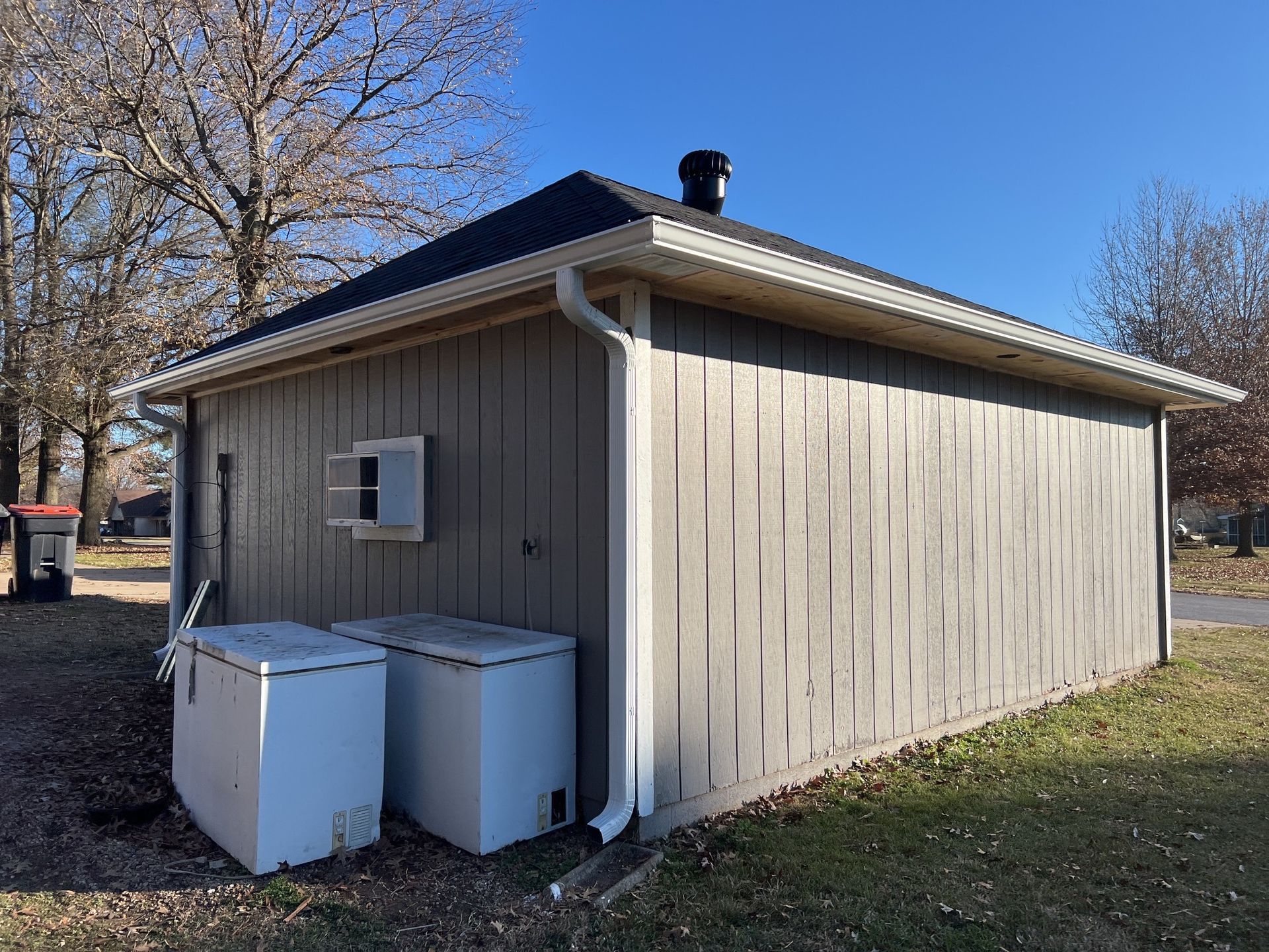 Two white chest freezers sit outdoors against the side of a gray, vertical-paneled shed with a dark roof and a drainpipe.