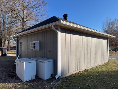 A gray shed with a black roof and white trim sits on grass, with two white chest freezers parked against the side wall.