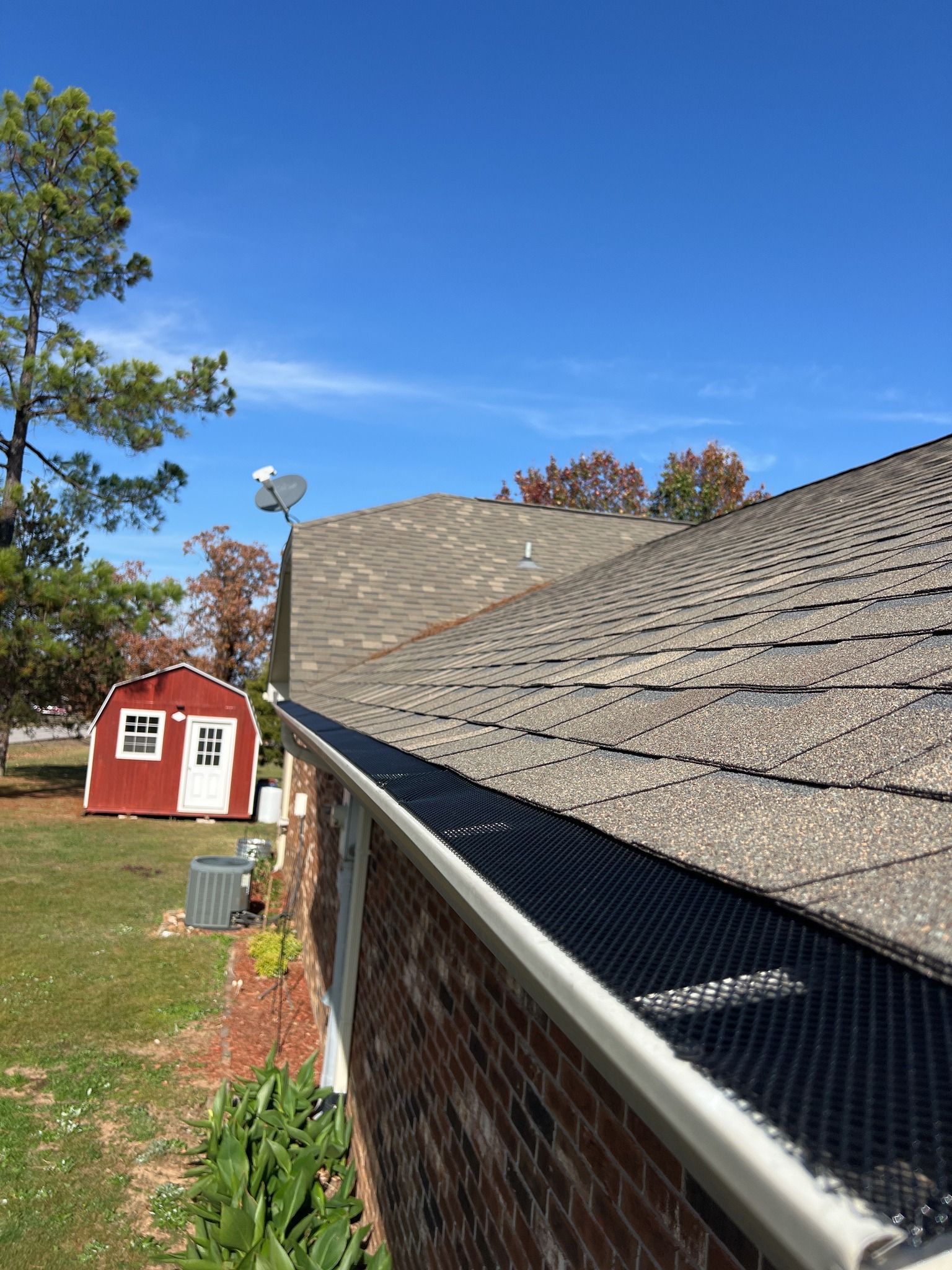 A roof edge with black mesh gutter guards, a brick exterior wall, a red shed in the yard, and a clear blue sky.