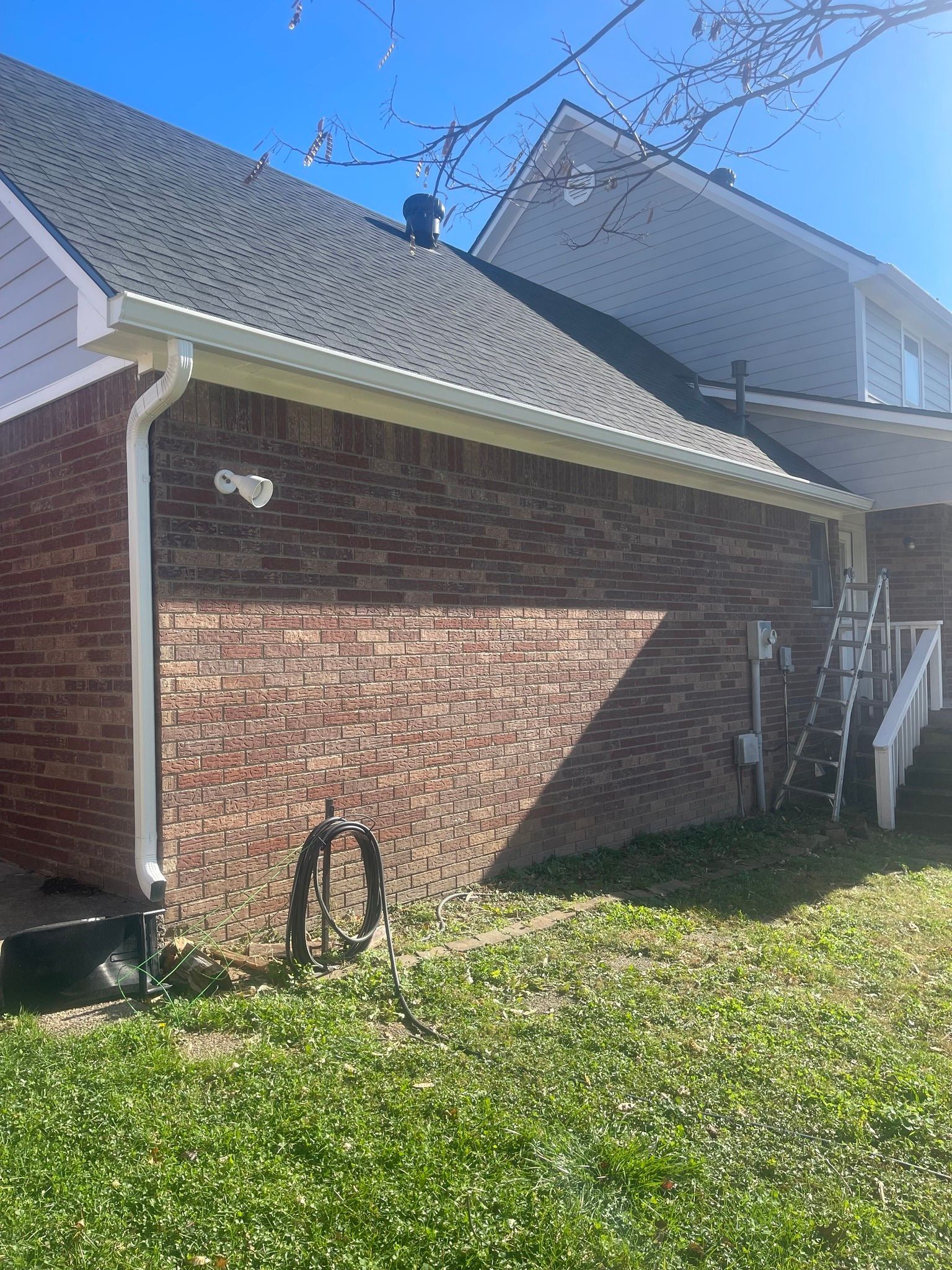 A side view of a residential brick house featuring a dark roof, white guttering, and a hose coiled on the lawn.