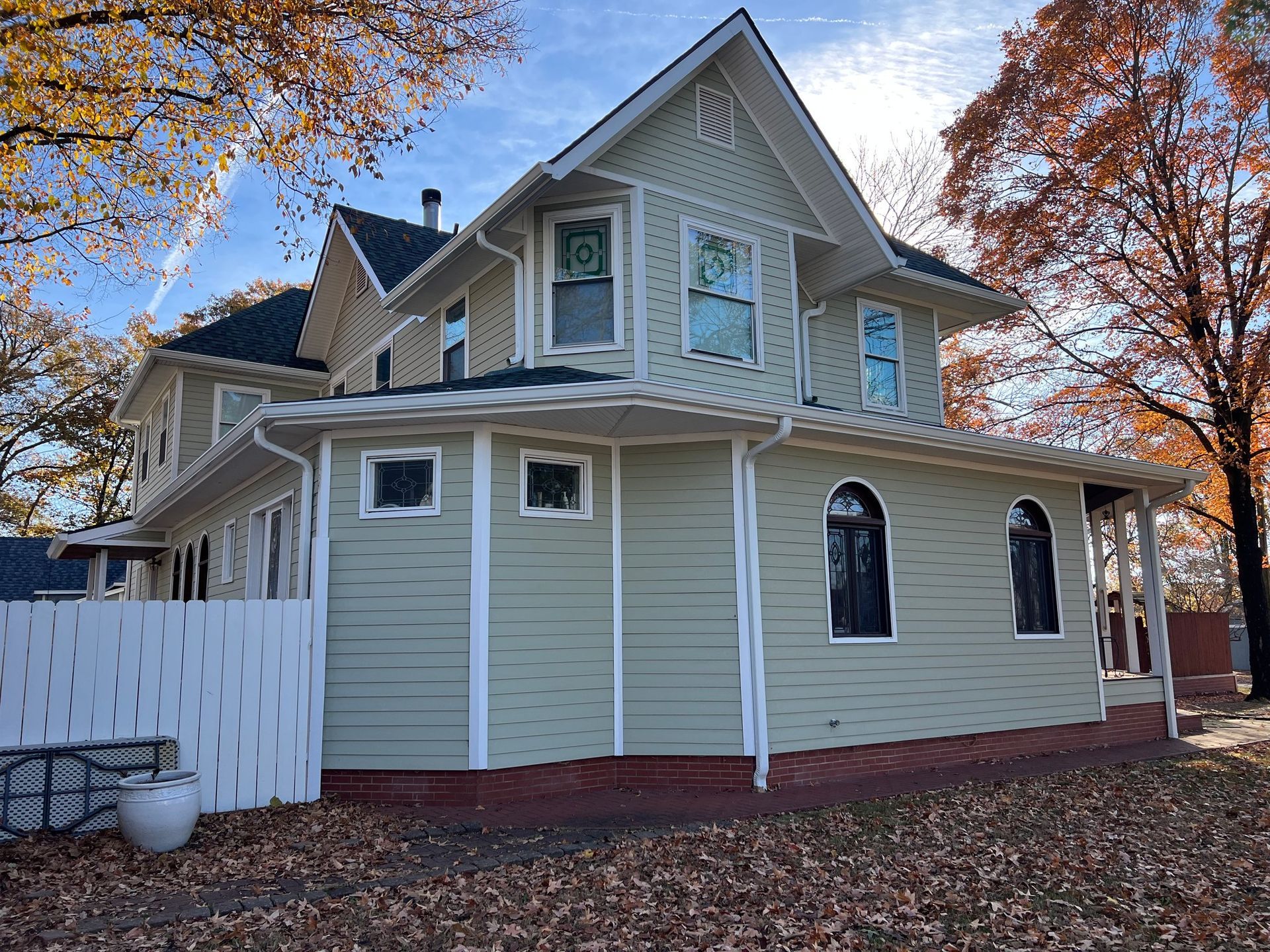 A light green two-story Victorian house with white trim and arched windows stands amidst autumn leaves under a blue sky.