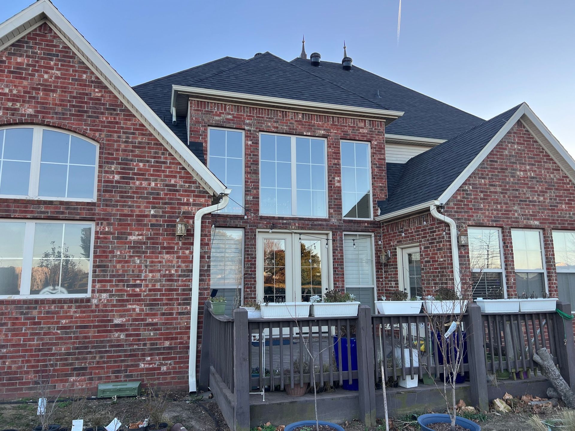 Back exterior of a two-story red brick house with dark roof shingles, large windows, and a dark wooden deck.