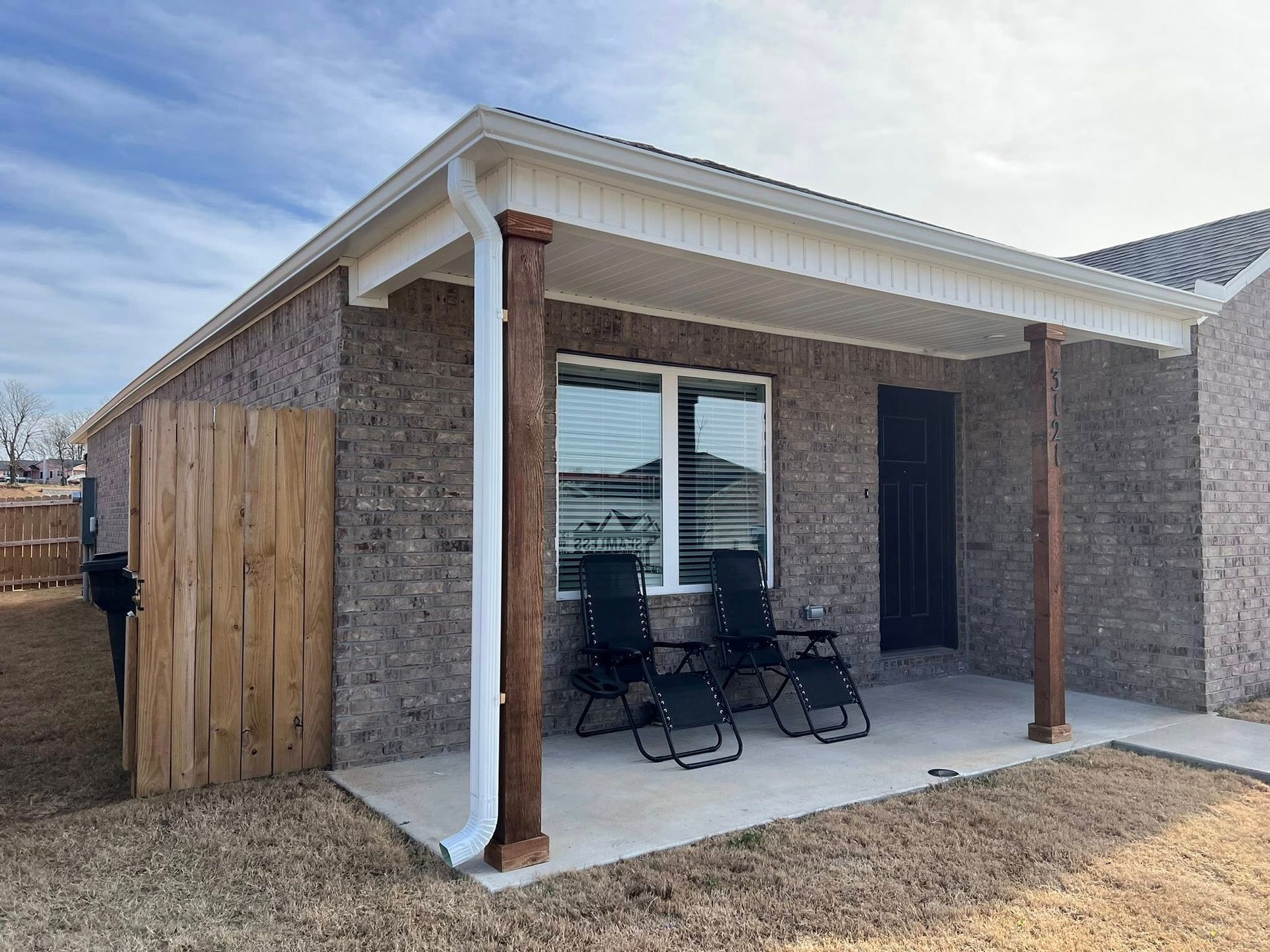 A brick house exterior with a covered concrete patio featuring two dark folding chairs, wooden posts, and a white downspout.