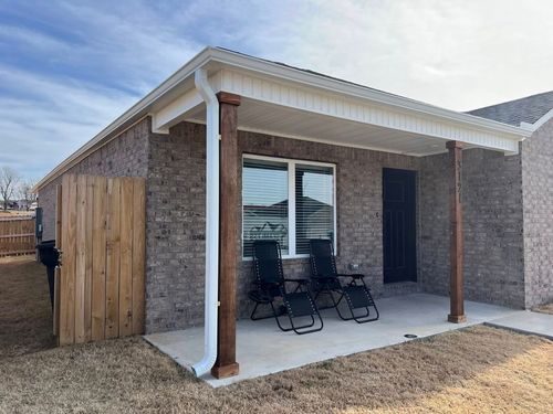 A grey brick house exterior with a covered concrete patio, two black lounge chairs, and wooden support columns.