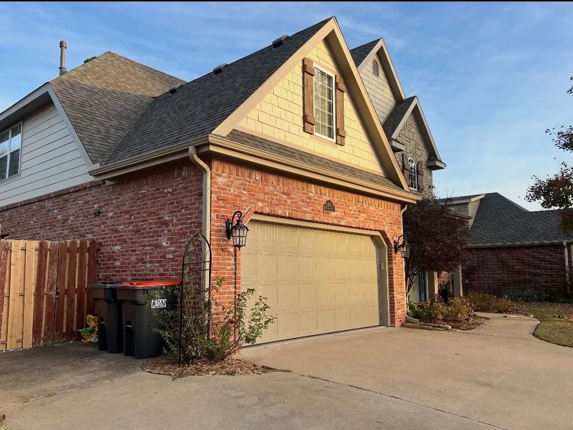 A two-story brick and siding house with a tan garage door, shingled roof, and a wooden fence on a sunny day.