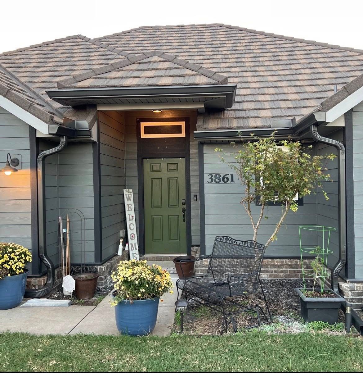 The front entrance of a grey house with a green door, potted plants, a decorative sign, and a metal bench.