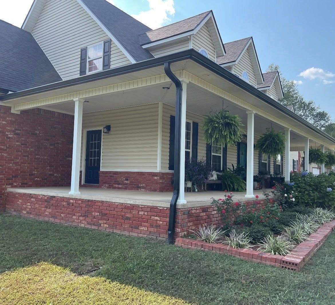 A wrap-around porch on a house with yellow siding and red brick accents, surrounded by a landscaped garden.