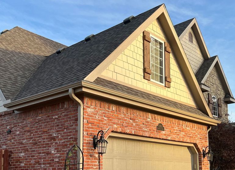 A two-story house with a brick ground floor, beige shingled gable, a dark shingled roof, and a garage under a blue sky.