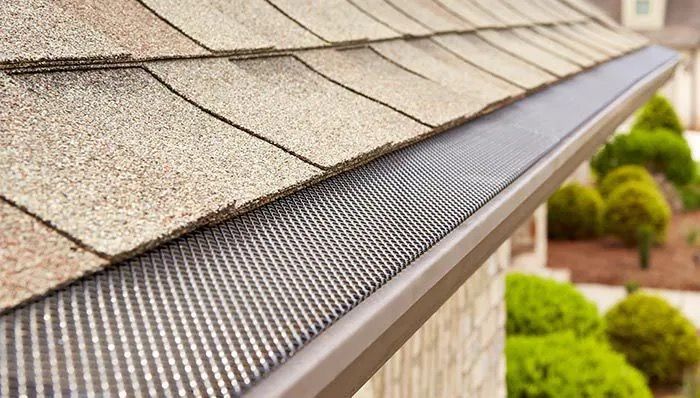 A black gutter guard installed on a house roof with brown shingles, viewed from a close-up, low-angle perspective.