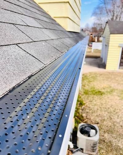 A perforated black gutter guard installed on a residential roof, with a yard and an air conditioning unit visible below.