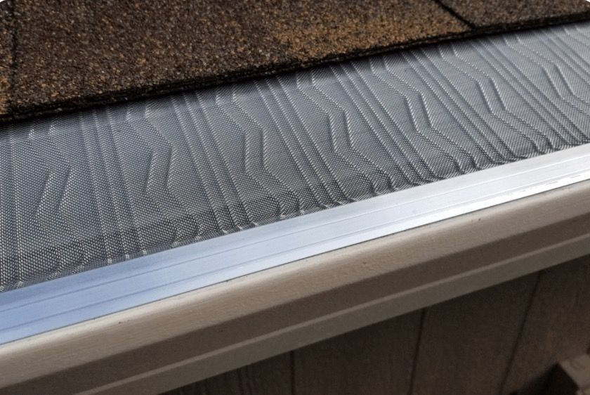 A silver gutter guard with a raised geometric pattern installed over a beige gutter and beneath brown roof shingles.