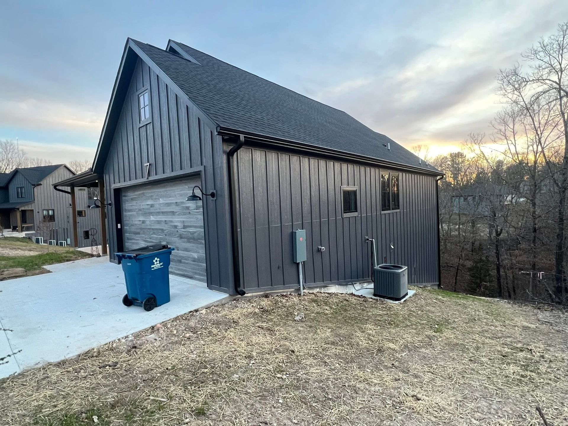 A modern dark-gray metal-sided detached garage with a gabled roof, a closed garage door, and a blue trash bin outside.