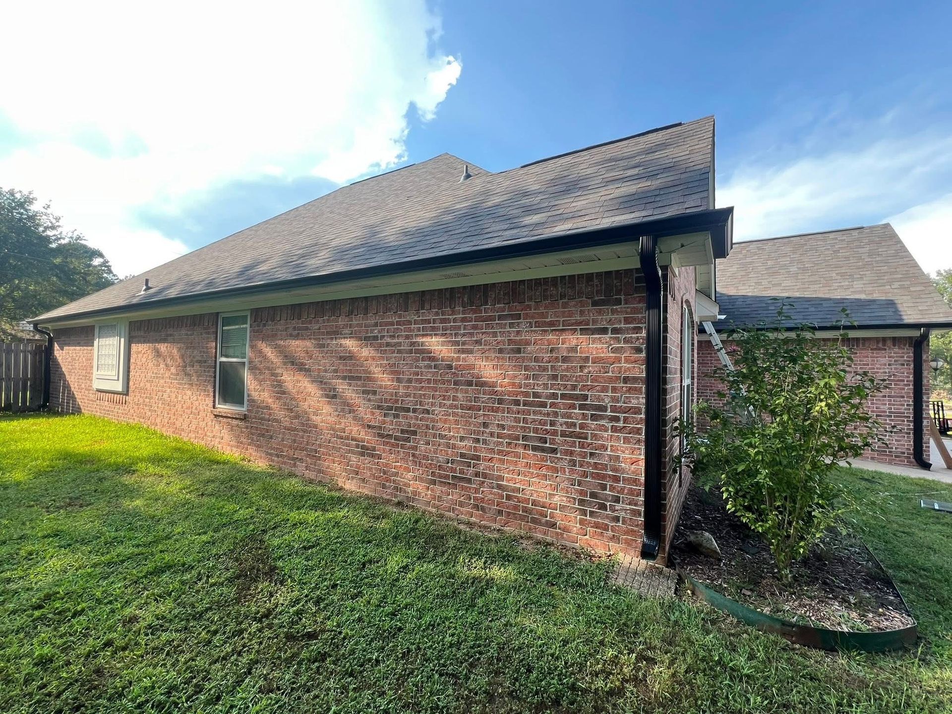 A side view of a residential brick house with a dark shingle roof, green lawn, and a small tree near the corner.