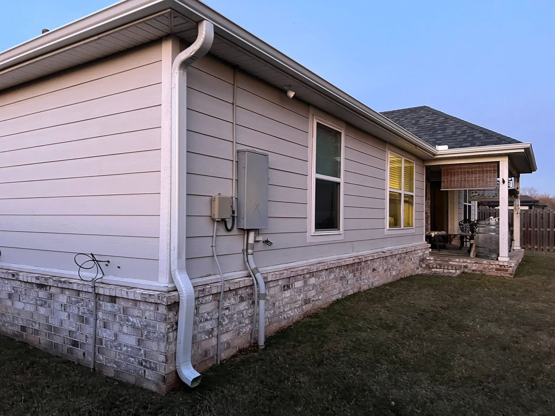 Exterior view of a house corner with light-colored horizontal siding, a stone base, a white downspout, and an electrical box.