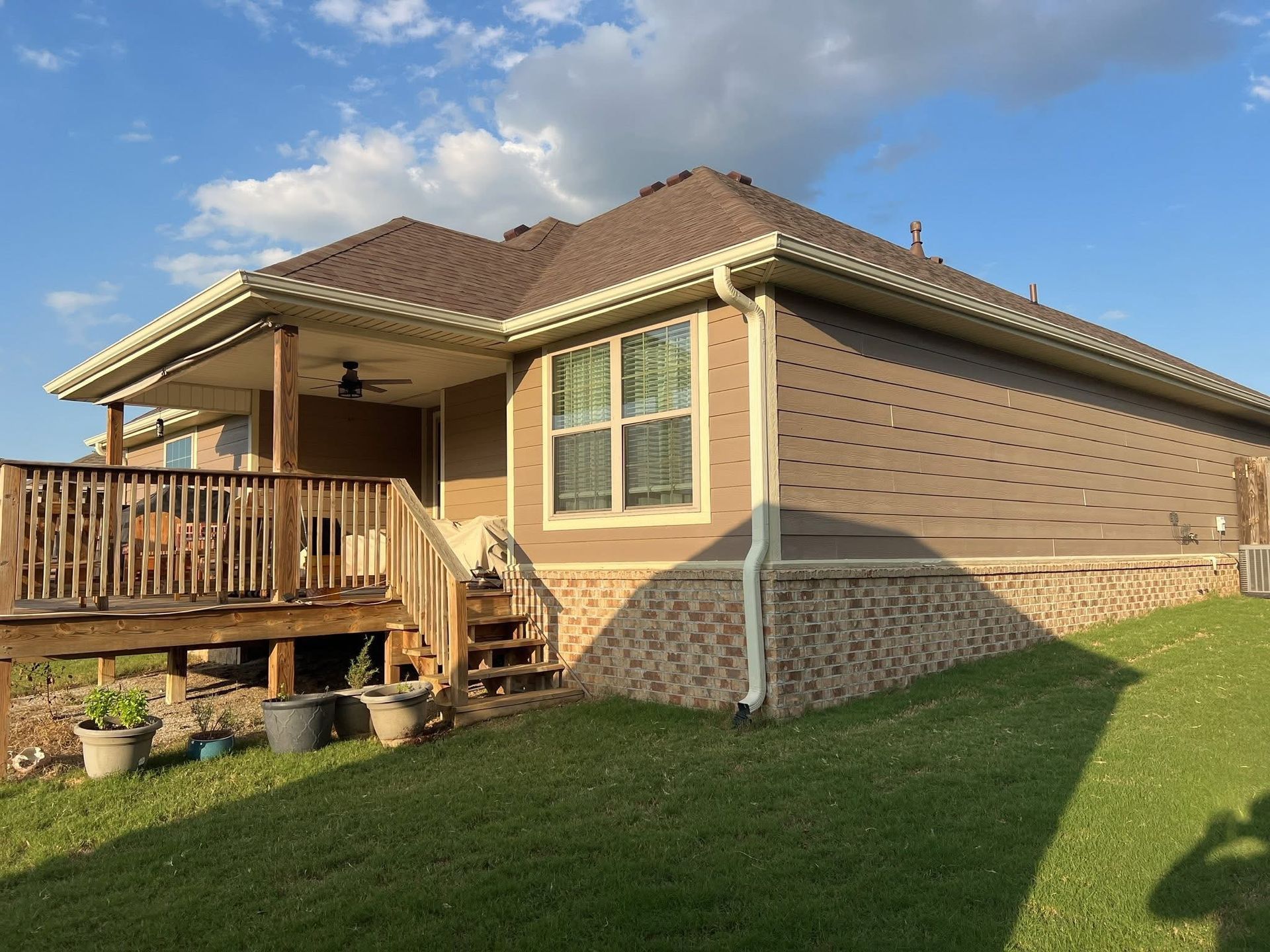 Side view of a one-story tan house with a brick foundation, brown siding, and a wooden deck under a bright blue sky.