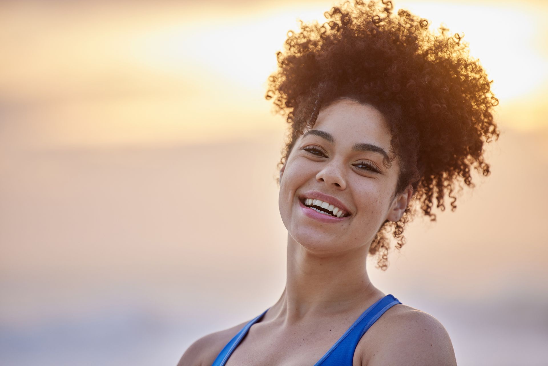 Woman smiles at the camera with a beach setting, wearing a blue sports bra, with natural, curly hair, against an orange sky.