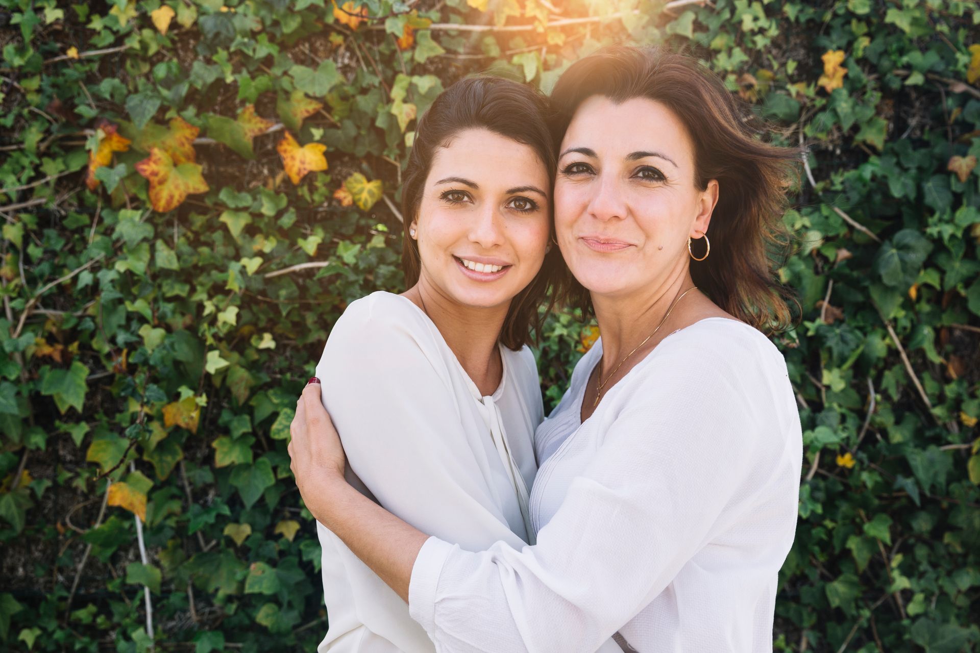 Two people in white shirts embrace each other in front of a green ivy wall with warm sunlight from above.