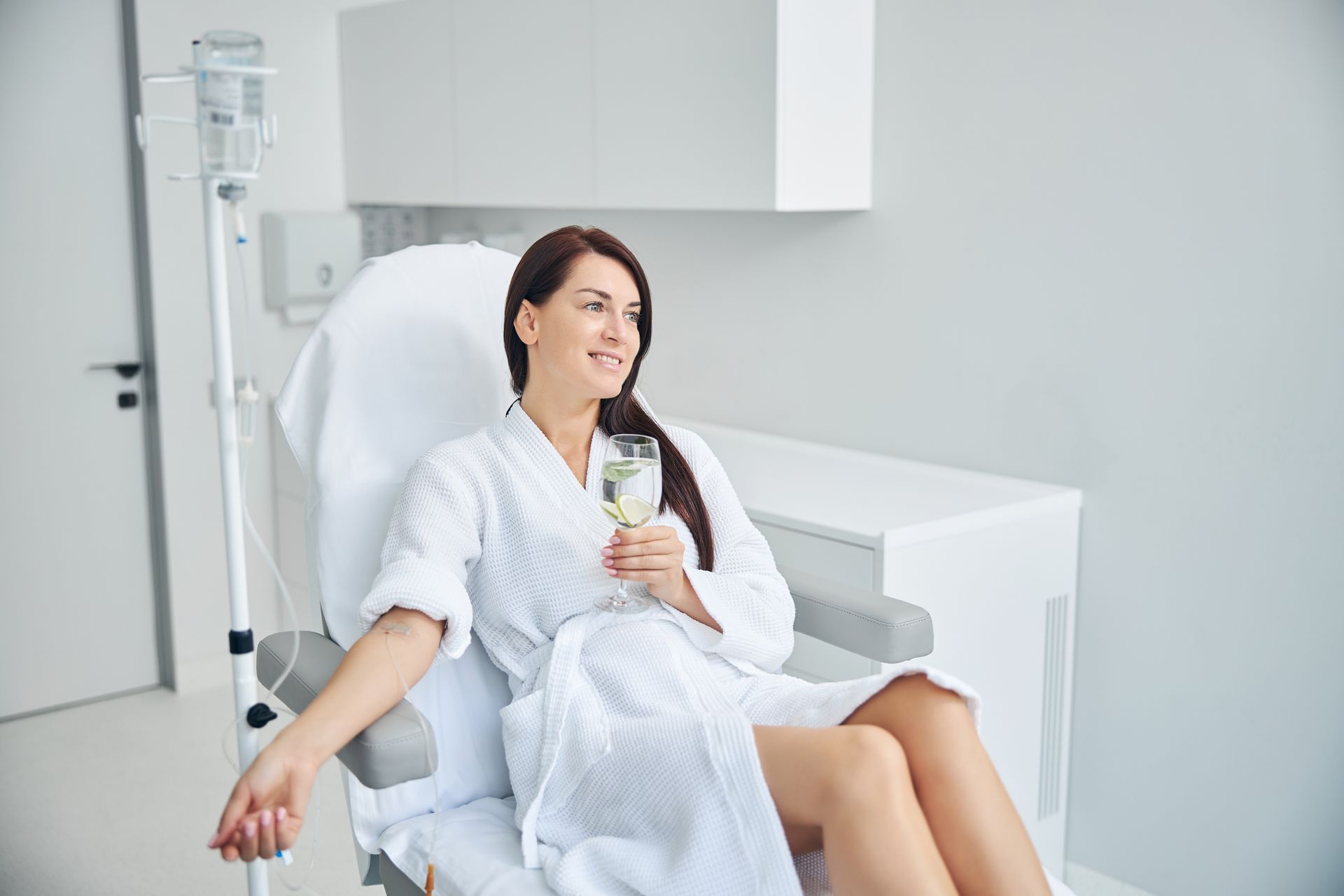 A smiling person in a white robe holds a glass while connected to an IV drip in a bright, modern clinic.