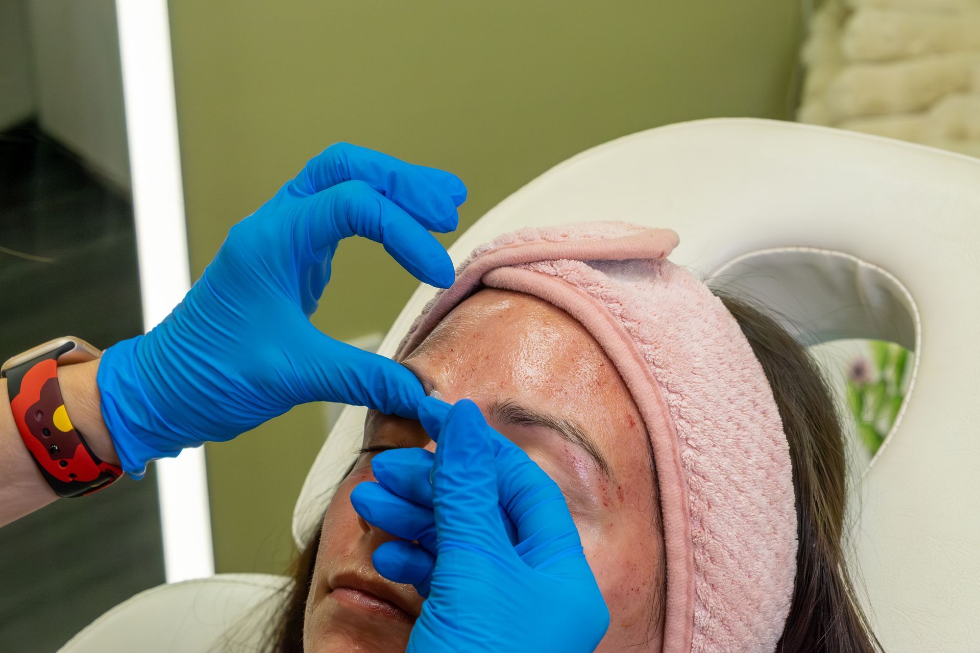 Gloved hands applying eye treatment to a person lying back with a pink headband in a clinic