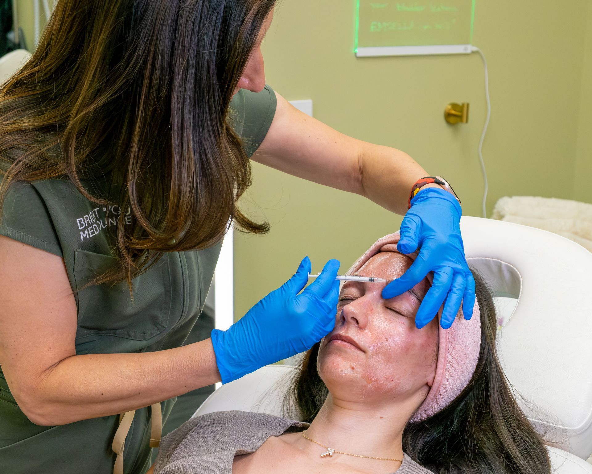 Esthetician applying facial treatment to a client with blue gloves and pink headband in a spa room