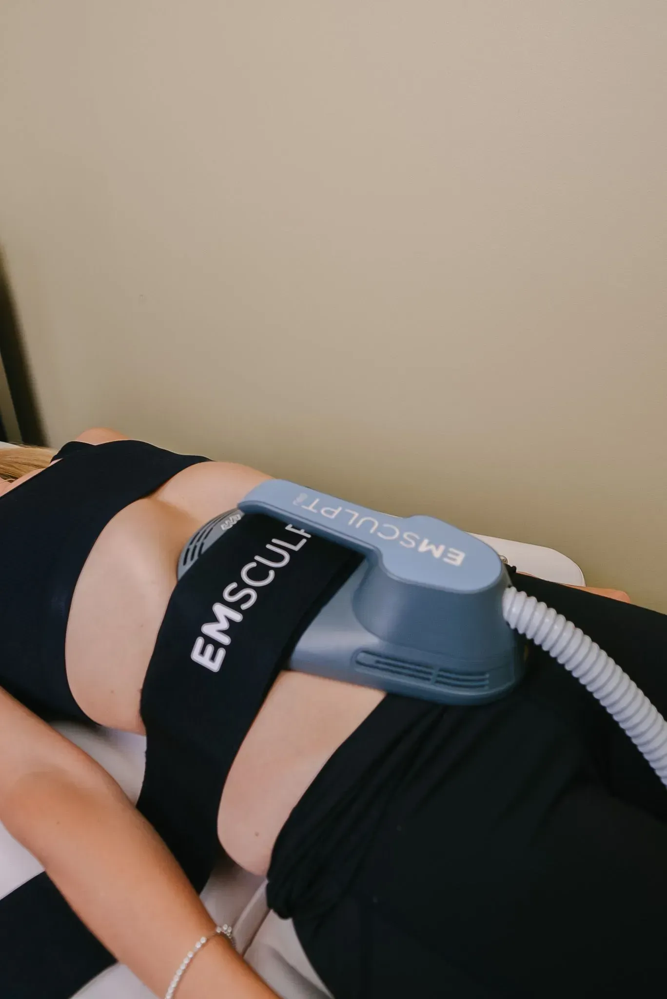 Woman lying down with an EMSCULPT device strapped to her abdomen. Black outfit, cream sheets, beige wall.