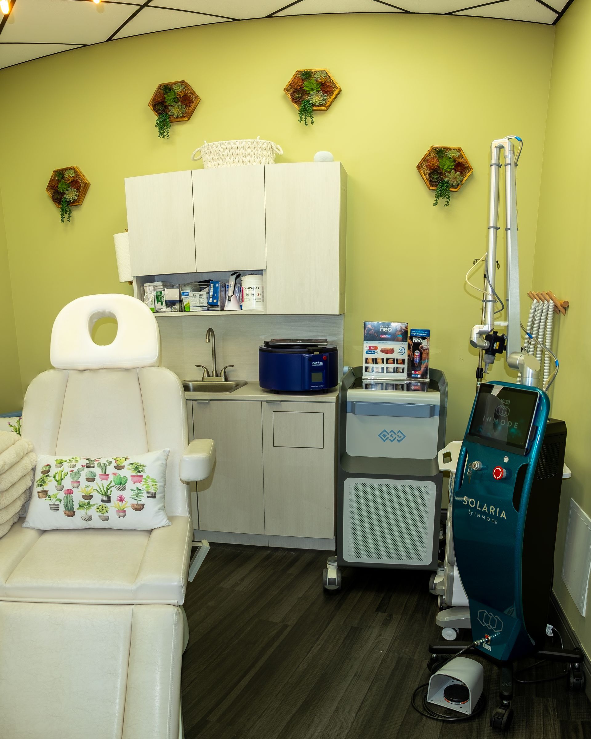 Spa treatment room with medical equipment, cream-colored chair, pale green walls, floral decor.