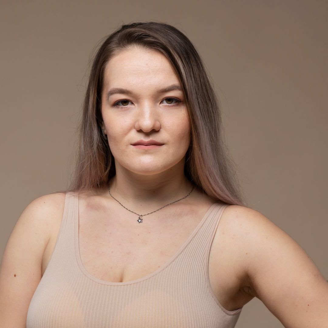 A woman in a tank top and necklace is standing in front of a brown background.