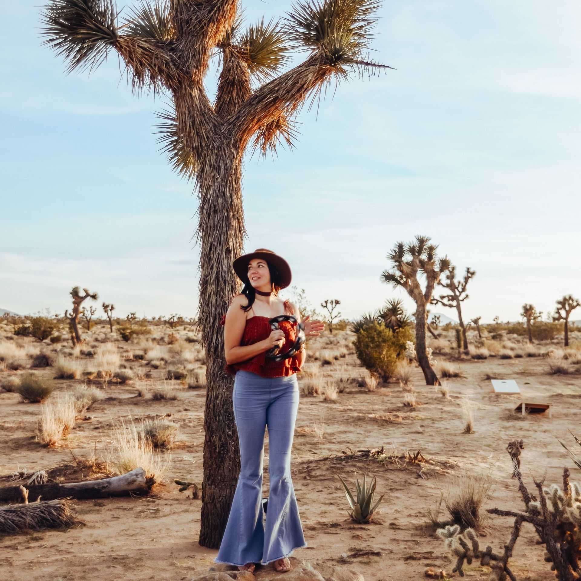 A woman in a hat is leaning against a tree in the desert.