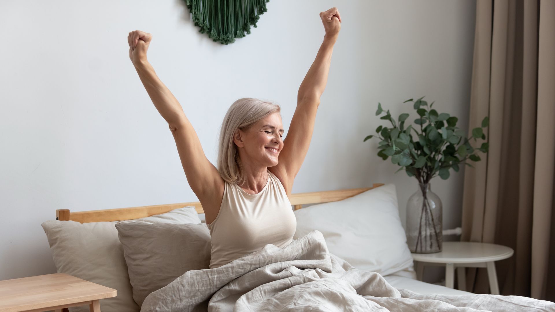 Woman sitting up in bed, arms raised in a stretch, smiling. Bedroom setting with plants and neutral colors.