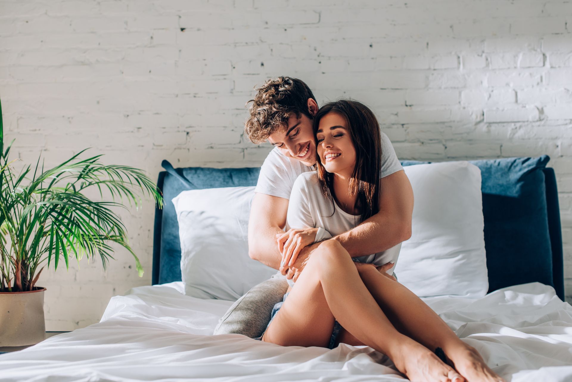 Couple embracing on a bed in a sunlit room, smiling. White bedding, a potted plant, and a blue headboard.