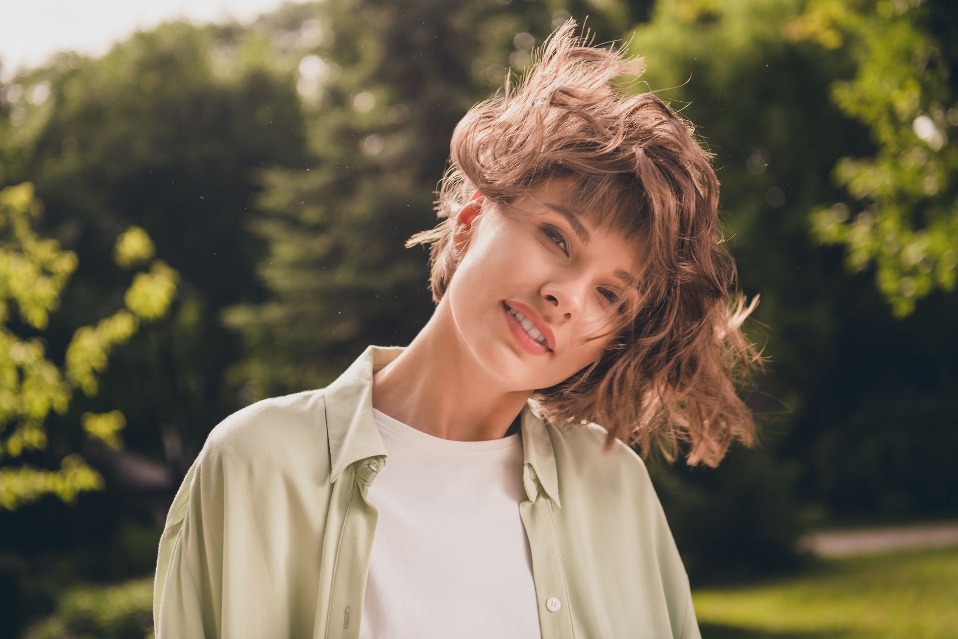 Woman with short, tousled hair smiles, wearing an unbuttoned green shirt and white shirt, outdoors with green trees.