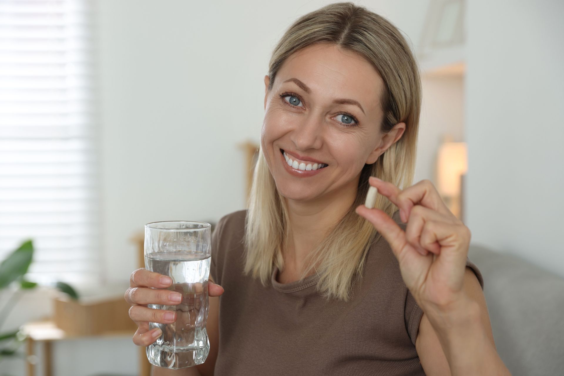 Woman holding a pill and a glass of water, smiling indoors.