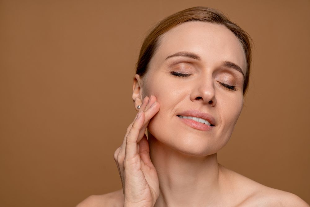 Woman with eyes closed, touching her cheek, against a brown background.
