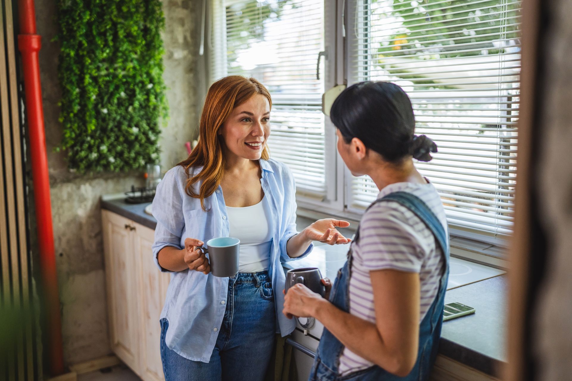 Two women talking in a kitchen, one holding a mug. The woman with red hair smiles.
