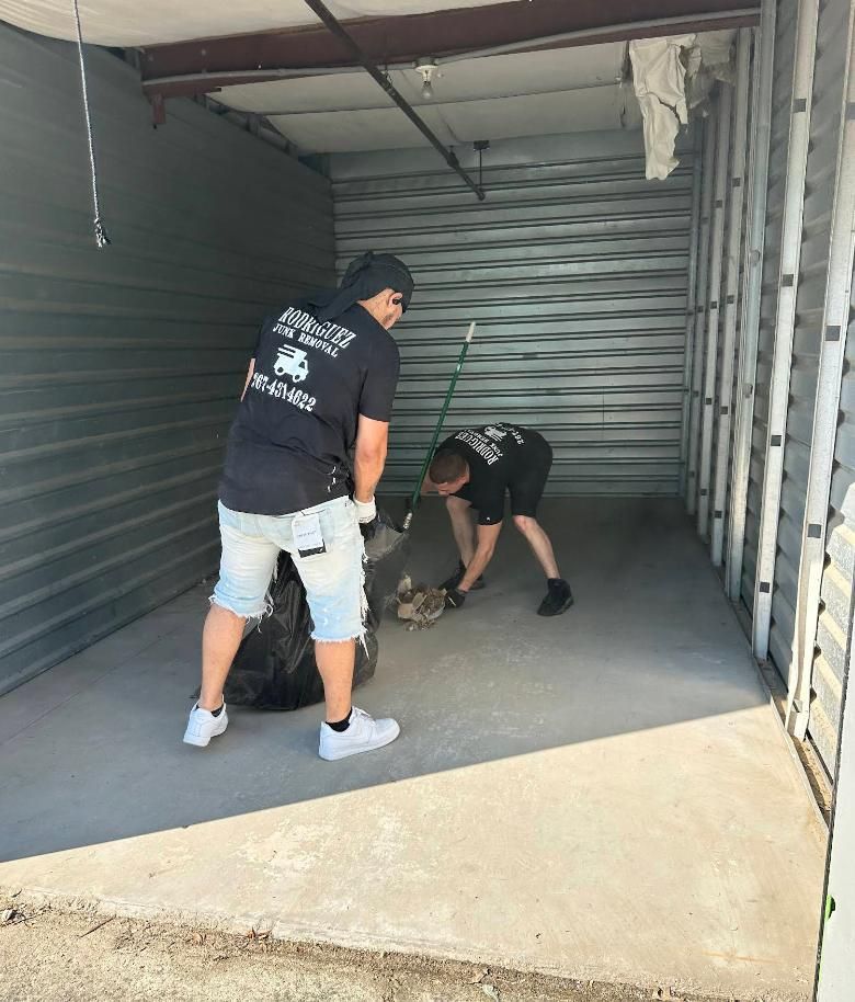 Two people in a storage unit cleaning up debris. One holds a trash bag, the other sweeps.