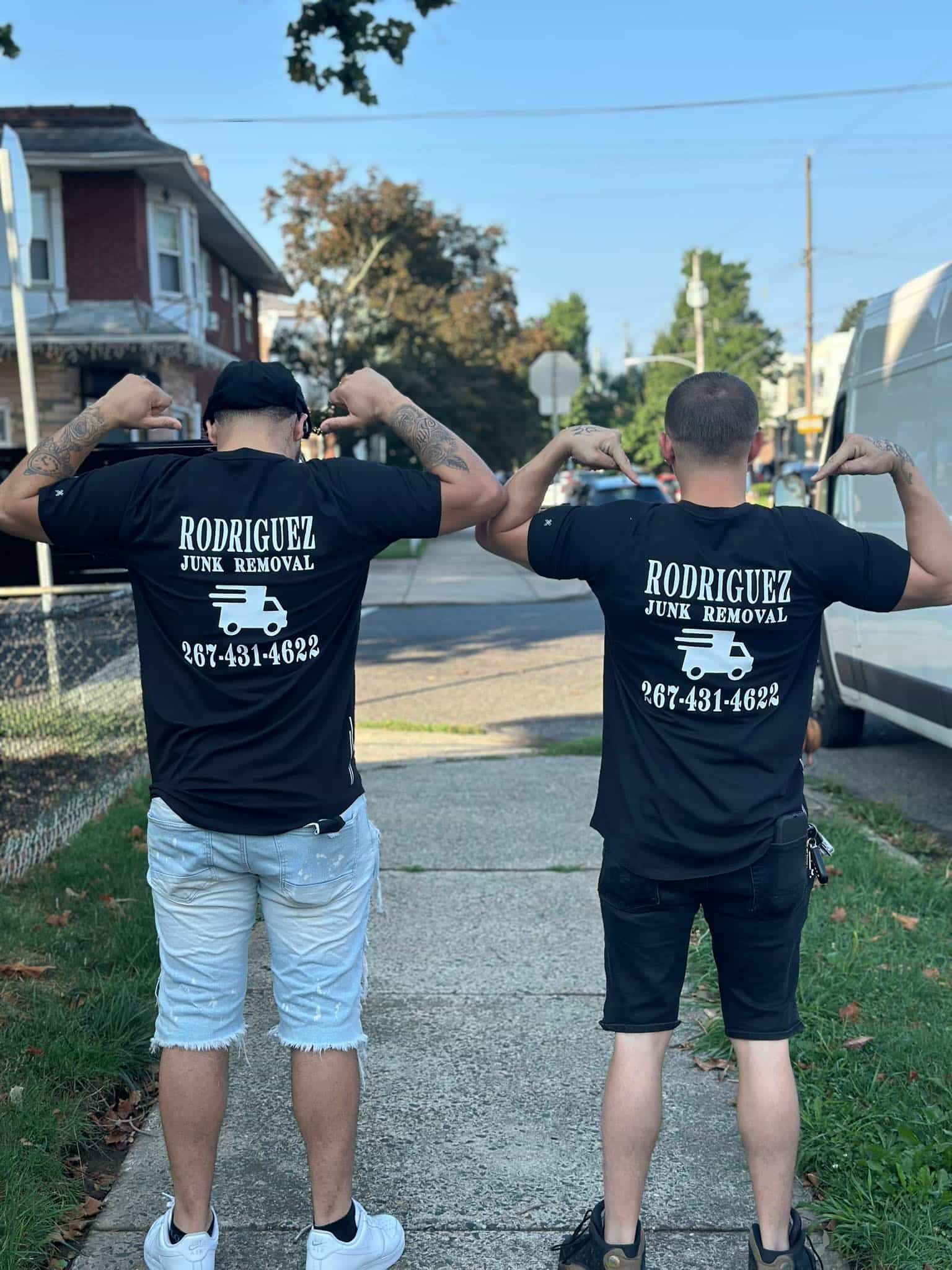 Two men in black shirts flexing, backs to the camera, outside.