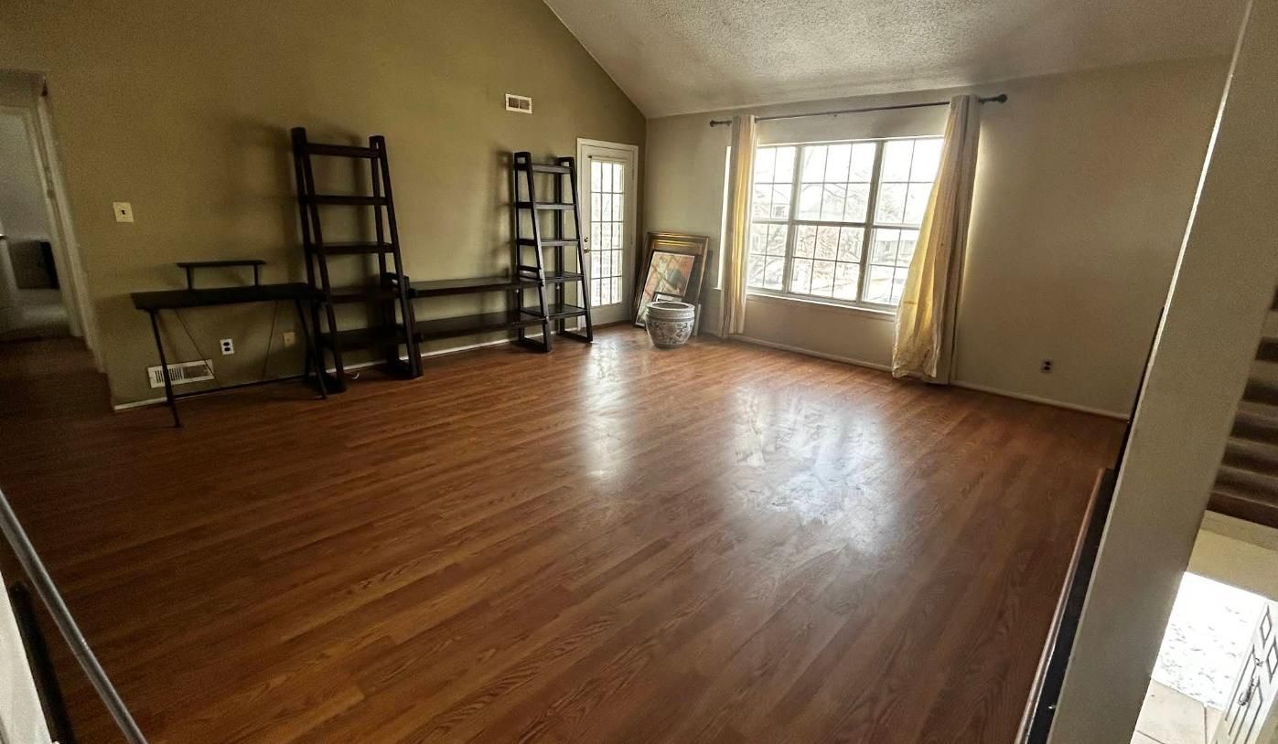 Empty living room with wood floors, shelves, a desk, and large window.